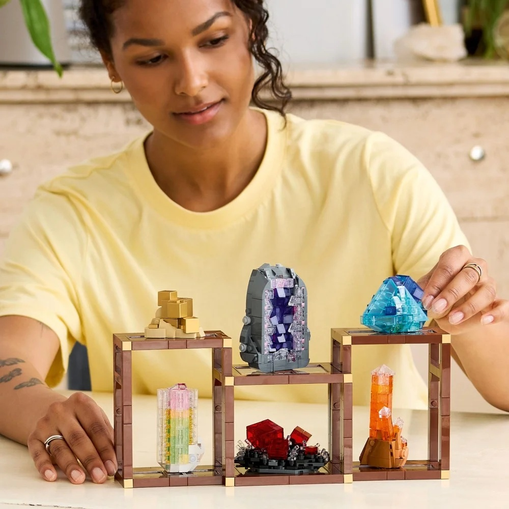 Kobieta przygląda się kryształom Lego na brązowych półkach. Wyświetlane są kryształy w różnych kolorach. Ma na sobie żółtą koszulę.