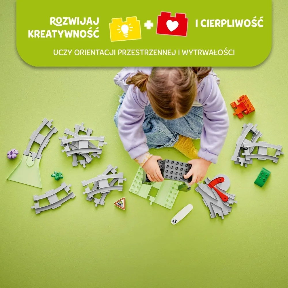 Dziecko bawi się zabawkami torami kolejowymi i klockami na zielonej powierzchni. Dziecko ma na sobie fioletową koszulę.