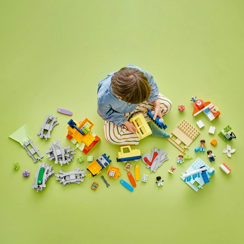 Dziecko siedzi otoczone kolorowymi klockami, w tym torami kolejowymi, pojazdami i figurkami, na zielonej powierzchni.