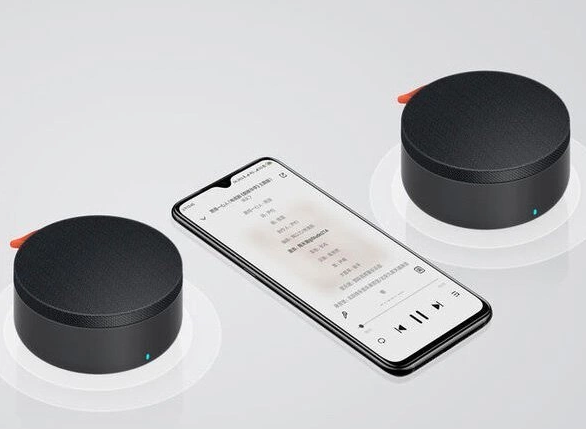 Dwa czarne głośniki z pomarańczowymi akcentami i smartfon wyświetlający elementy sterowania muzyką. Tło jest białe.