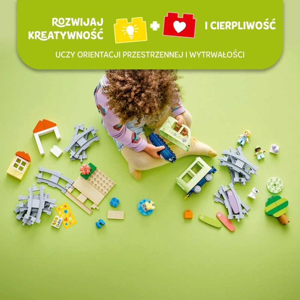 Dziecko bawi się kolorowymi częściami pociągu na zielonej powierzchni. Zabawki obejmują tory kolejowe, domki i figurki.