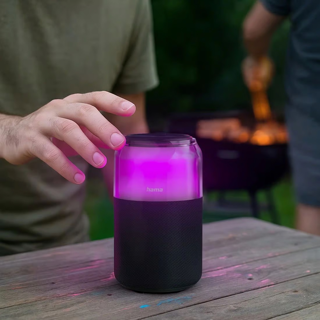 Dłoń dotyka świecącego na różowo głośnika na drewnianym stole. W tle grill i inna osoba.