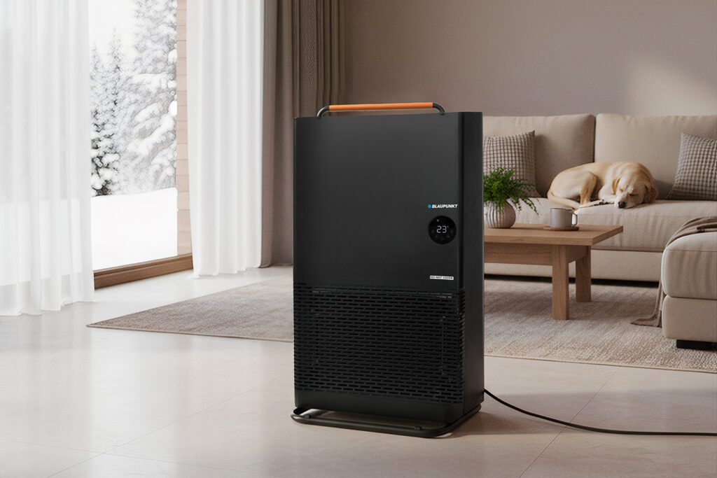 Czarny oczyszczacz powietrza w pokoju. Pies na kanapie. Okno ze śniegiem na zewnątrz. Beżowa sofa i dywan. Drewniany stół.