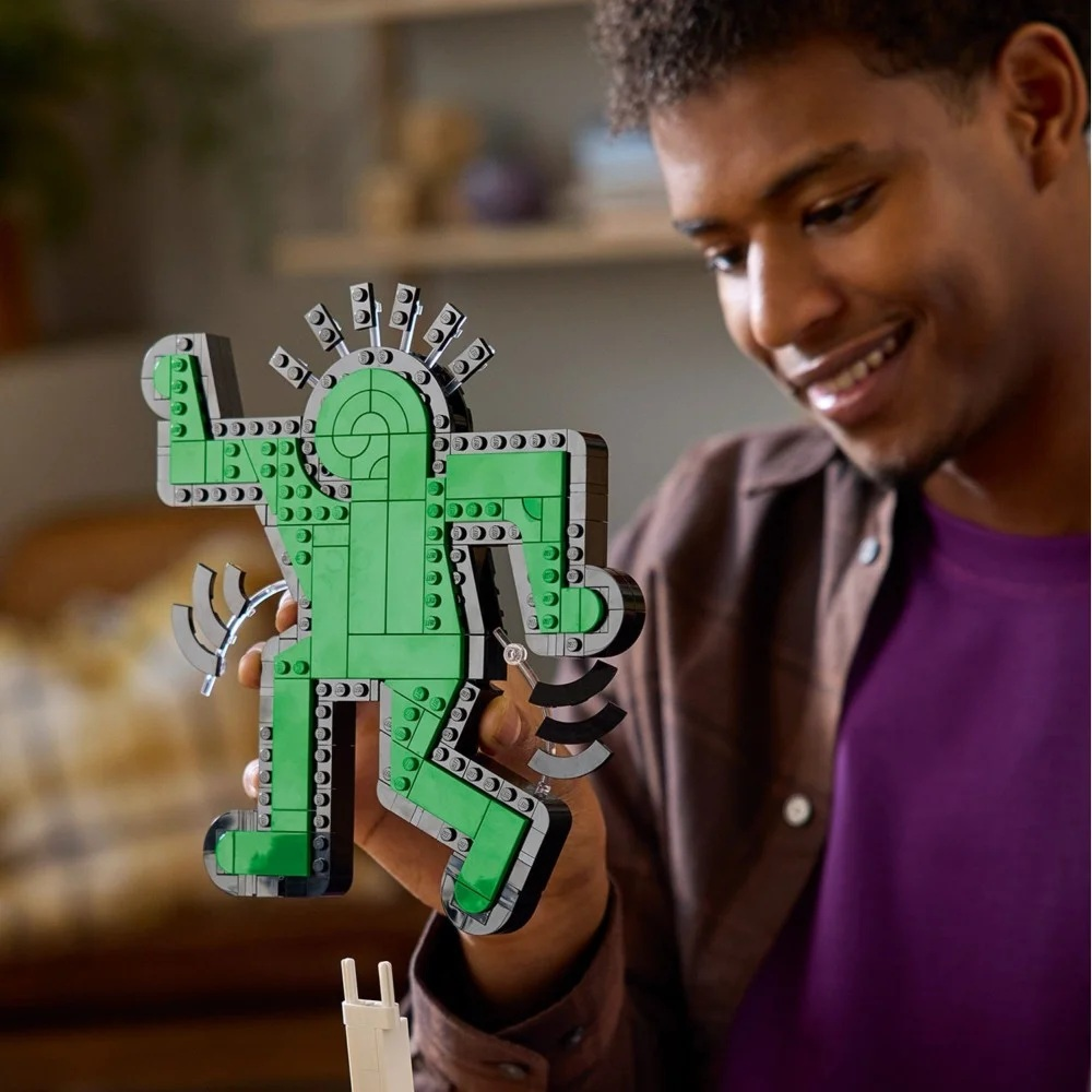 Mężczyzna trzyma zieloną figurkę Lego. Figurka to stylizowana osoba z czarnymi konturami. Mężczyzna uśmiecha się i ma na sobie fioletową koszulę.