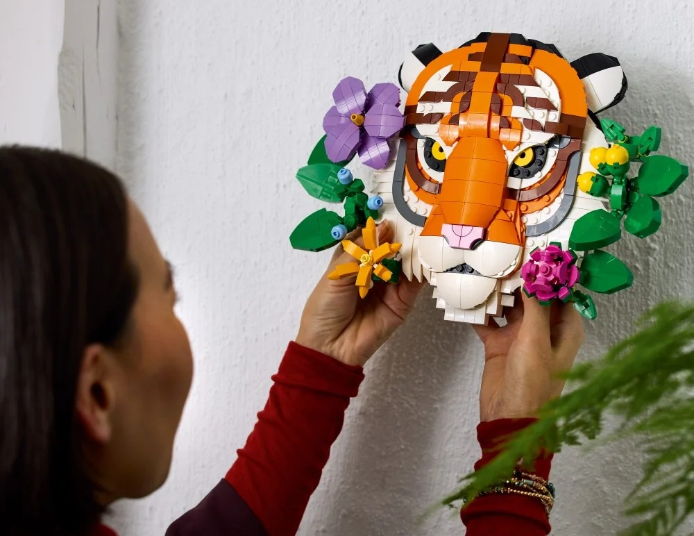 Kobieta trzyma głowę tygrysa Lego ozdobioną kwiatami. Tygrys jest pomarańczowy, biały i czarny. W tle biała ściana.