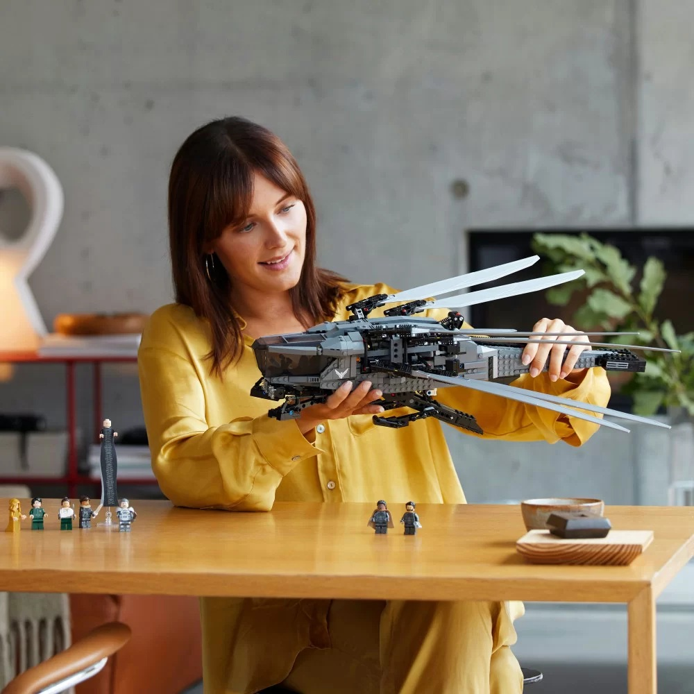 Kobieta w żółtej koszuli trzymająca statek kosmiczny Lego. Mini figurki i miska na drewnianym stole.