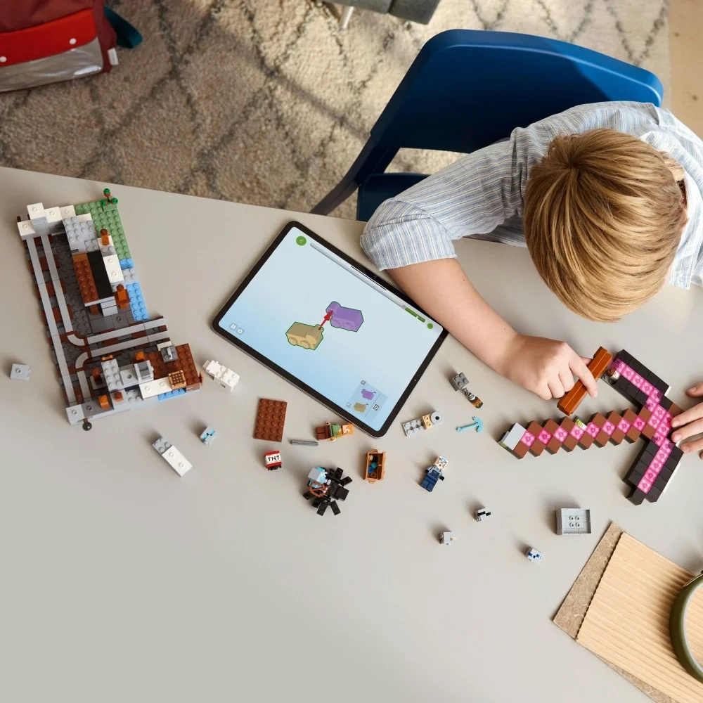 Dziecko buduje z Lego na stole, z iPadem pokazującym instrukcje. Widoczne są klocki Lego i kilof.