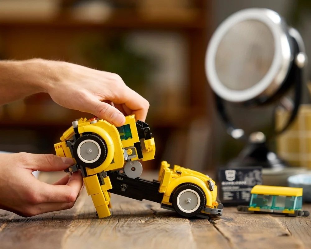 Ręce składające żółty samochód-zabawkę Lego Bumblebee na drewnianej powierzchni. Mikrofon i inne klocki Lego w tle.