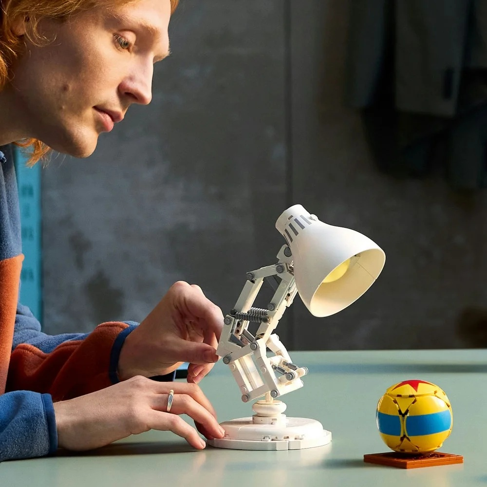 Osoba montuje białą lampę Lego obok piłki Pixar na stole. Osoba ma rude włosy i ma na sobie niebiesko-pomarańczową koszulę.
