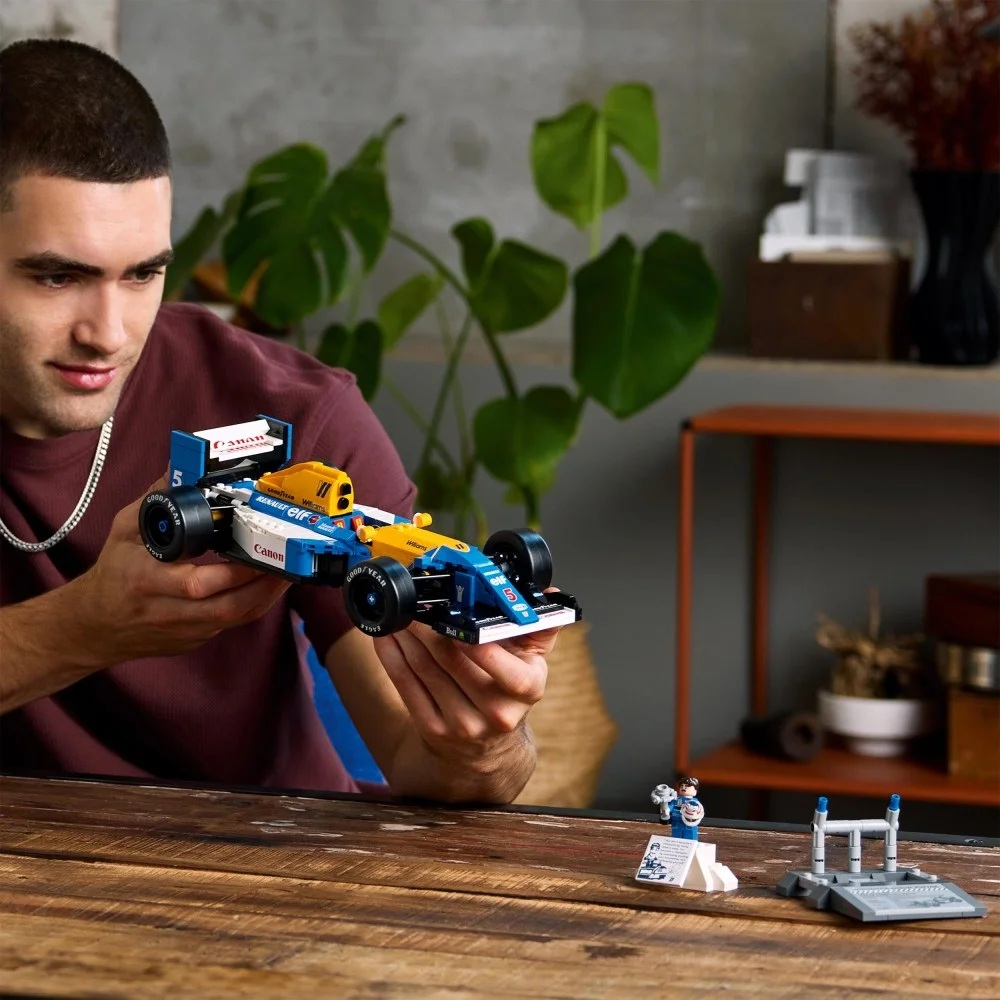 Mężczyzna trzyma niebiesko-żółty samochód wyścigowy Lego. Mały model samochodu wyścigowego i stojak znajdują się na drewnianym stole.