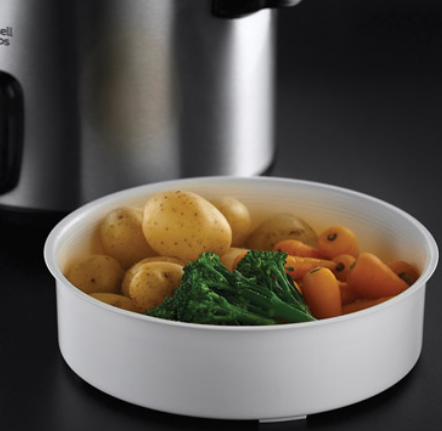 Biała miska zawiera ziemniaki, marchewkę i brokuły. Srebrne urządzenie znajduje się w tle. Jedzenie leży na czarnej powierzchni.