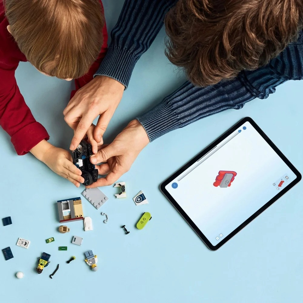 Dziecko i dorosły składają zestaw Lego. Tablet wyświetla instrukcje. Klocki i figurki Lego rozrzucone na niebieskiej powierzchni.