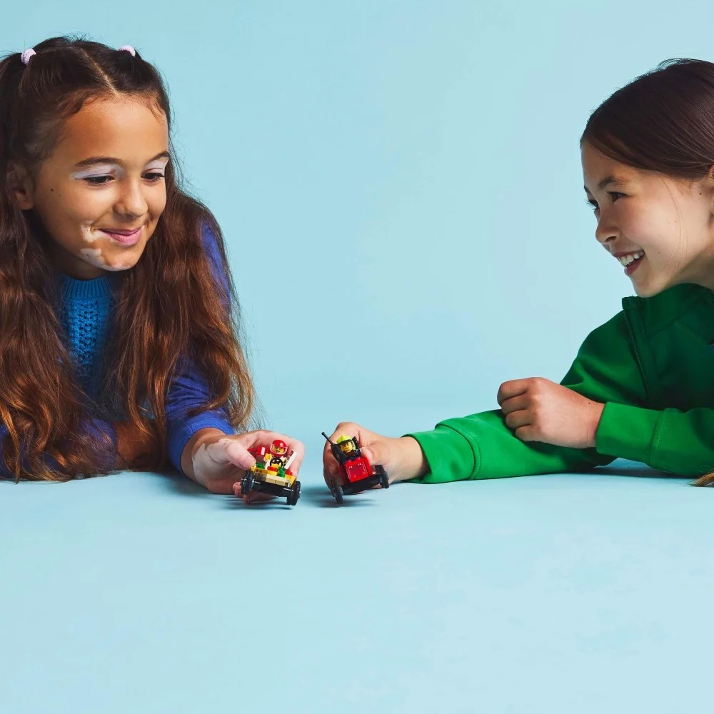 Dwie dziewczyny bawią się samochodzikami na niebieskim tle. Jedna ma na sobie niebieski, druga zielony. Obie się uśmiechają.