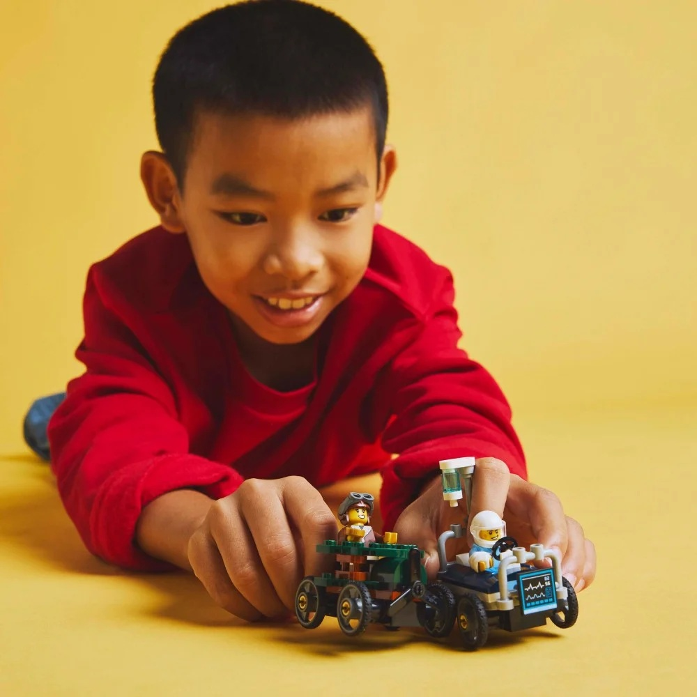 Chłopiec w czerwonej koszuli bawi się samochodzikami Lego na żółtej powierzchni. Samochody mają figurki Lego. Chłopiec się uśmiecha.