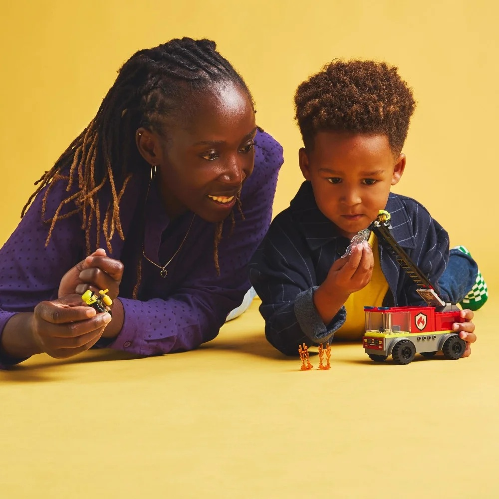 Kobieta i dziecko bawią się zabawkami w postaci wozów strażackich Lego. Leżą na żółtej powierzchni. Kobieta trzyma małą figurkę Lego.