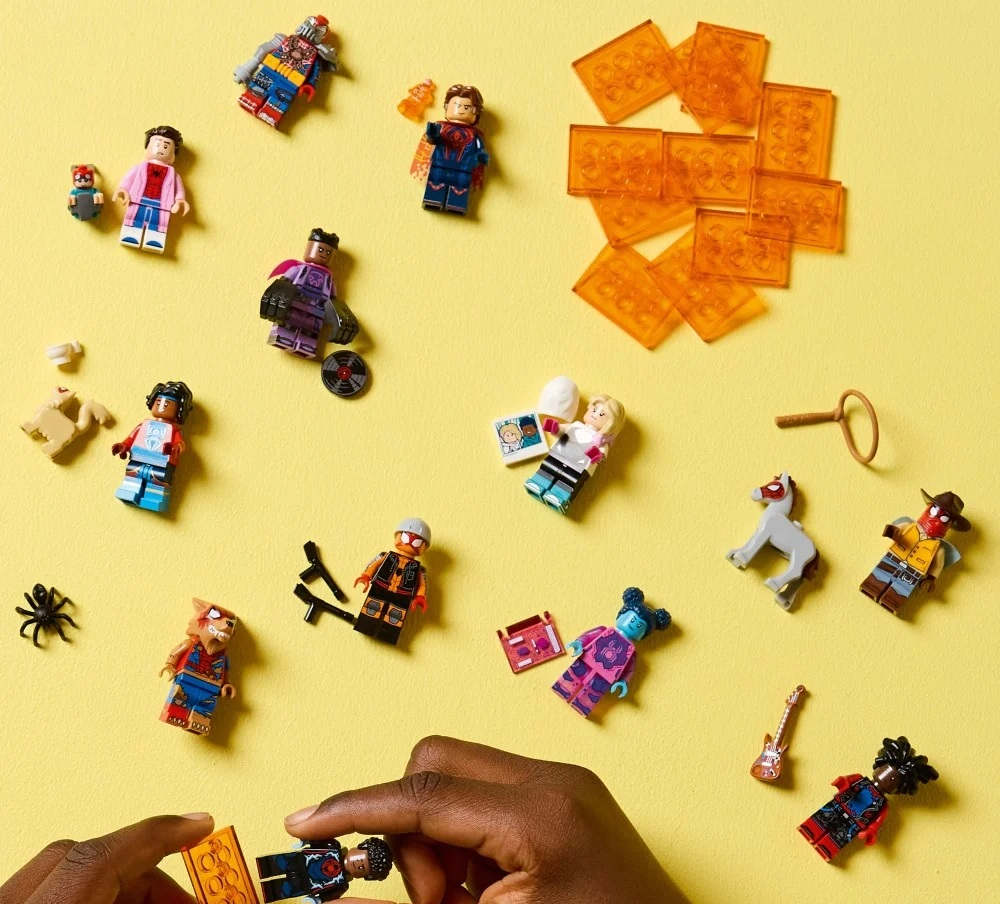 Figurki i akcesoria Lego na żółtej powierzchni. Ręka składa figurkę. Różne figurki są rozrzucone w tle.