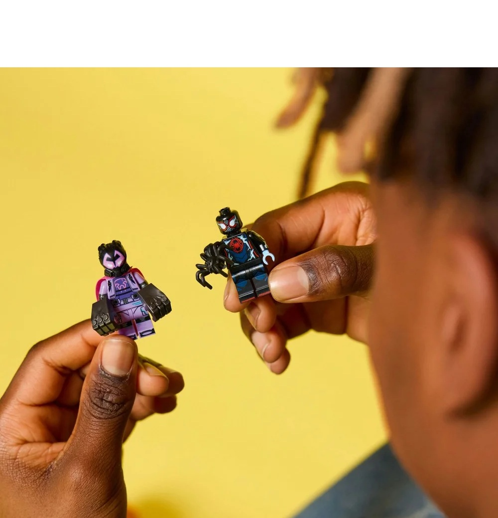 Osoba trzyma dwie figurki Lego Spider-Mana na żółtym tle. Jedna figurka jest fioletowa, druga czarna z czerwonym.