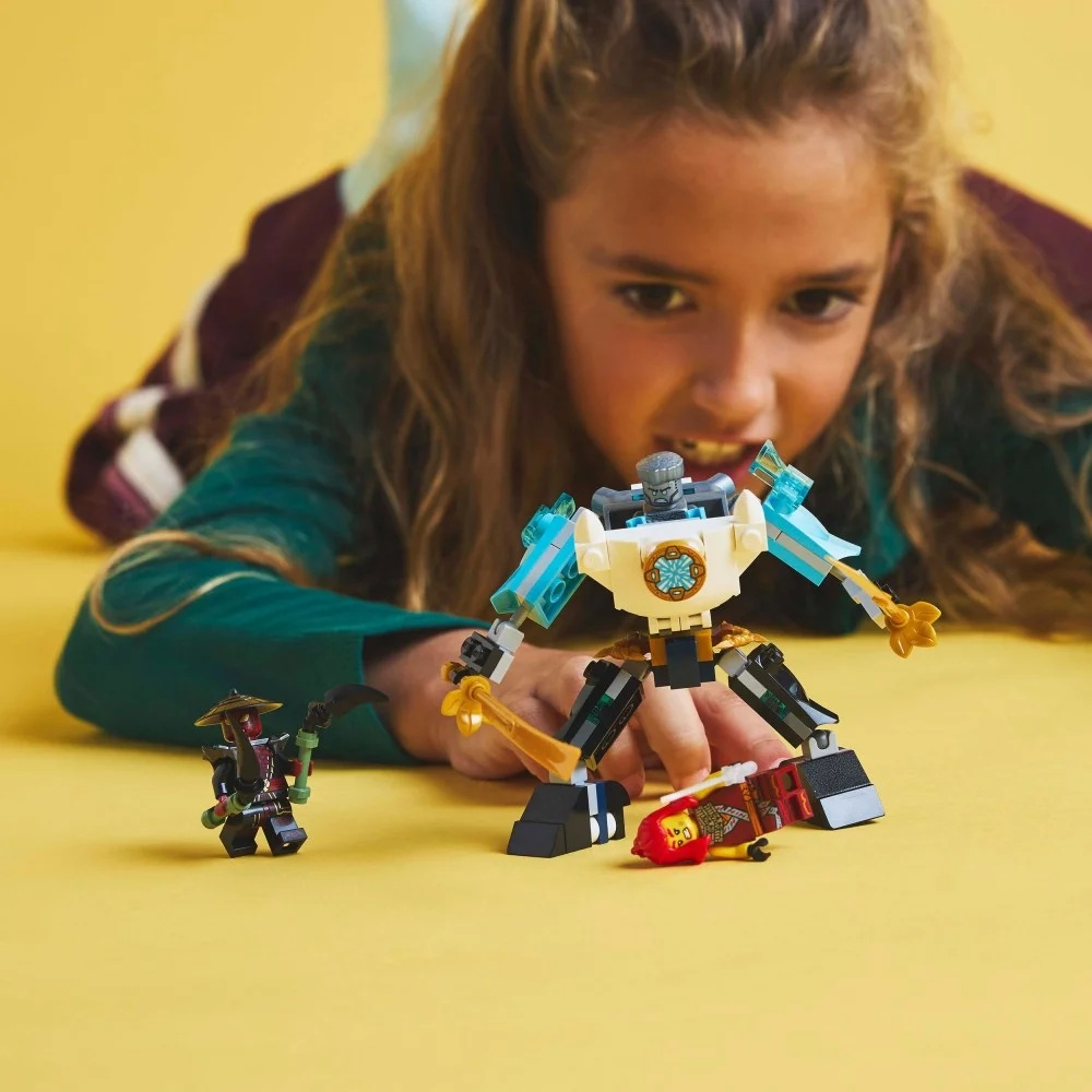 Dziewczyna bawi się figurkami Lego: robotem, ninja i upadłą postacią, wszystko na żółtej powierzchni.