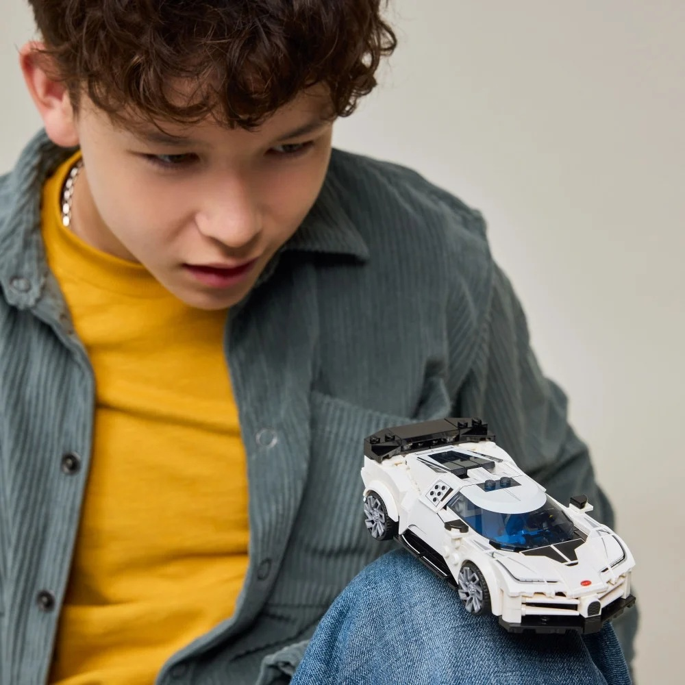 Chłopiec patrzy na biały samochodzik na kolanach. Ma na sobie żółtą koszulę, szarą kurtkę i niebieskie dżinsy.