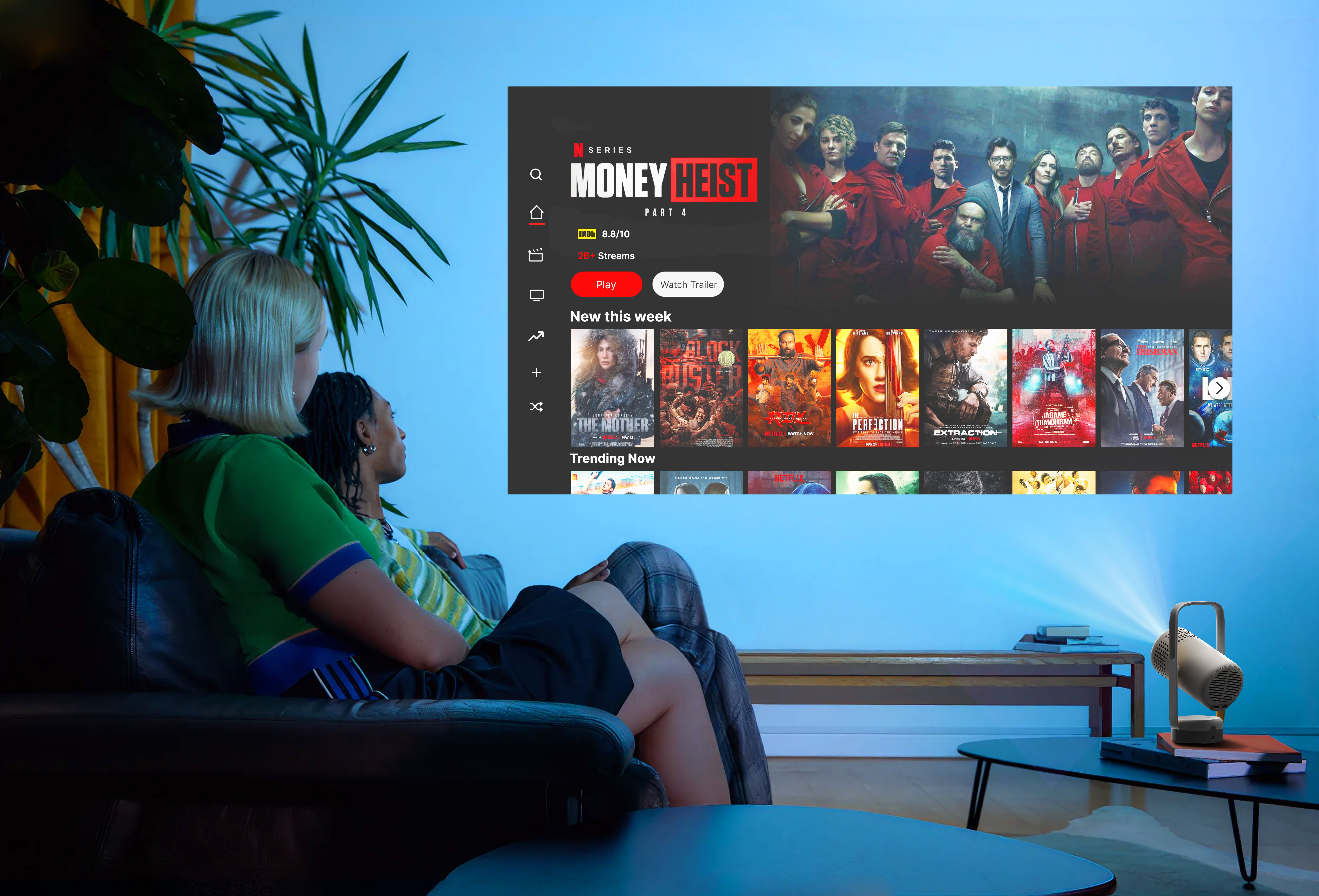 İki kadın ekranda film izliyor. Bir projektör masada. Film Money Heist.