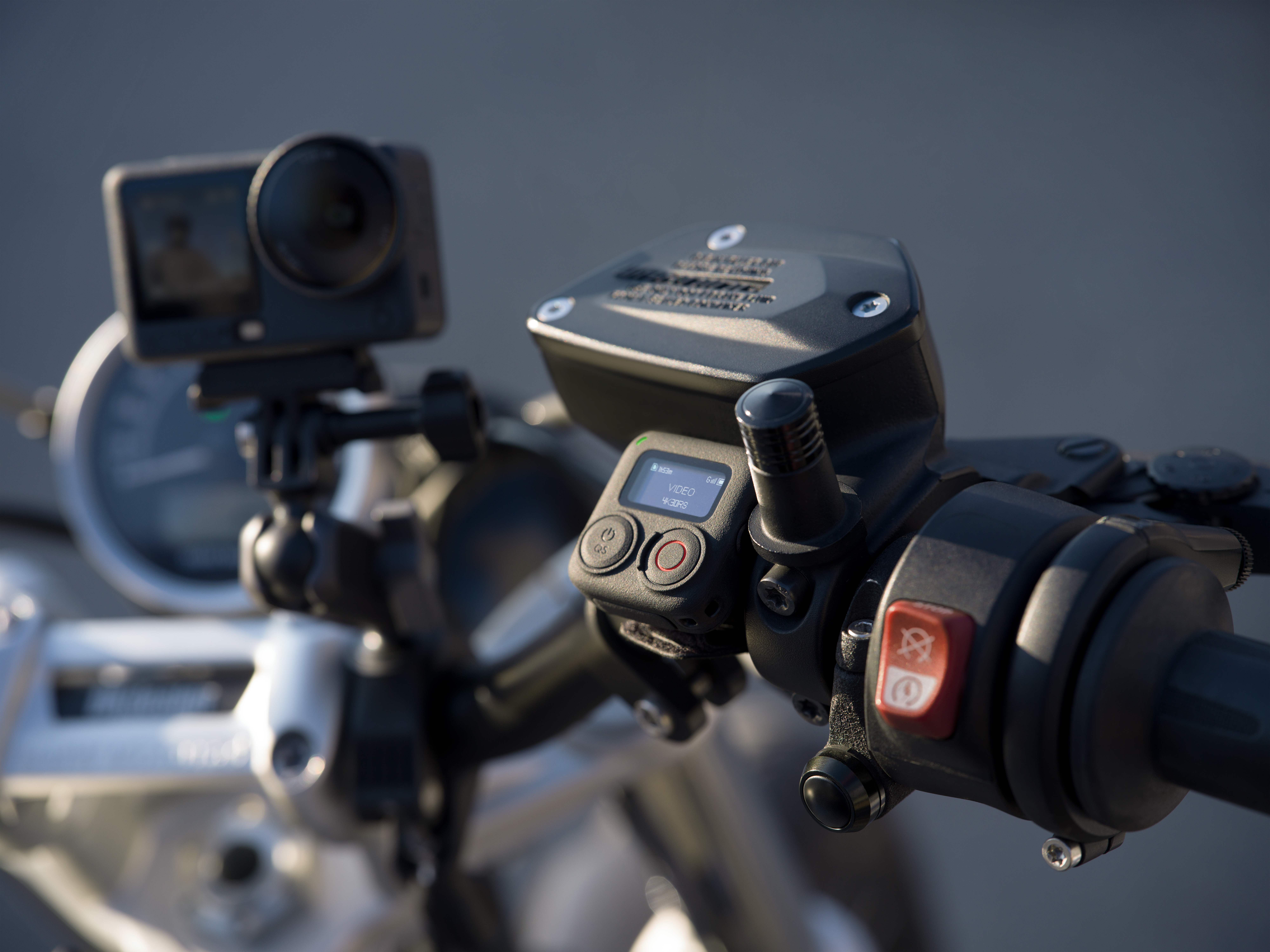 Kierownica motocykla z kamerą, przyciskami i wyświetlaczem. Czarne i czerwone detale na szarym tle, akcja.