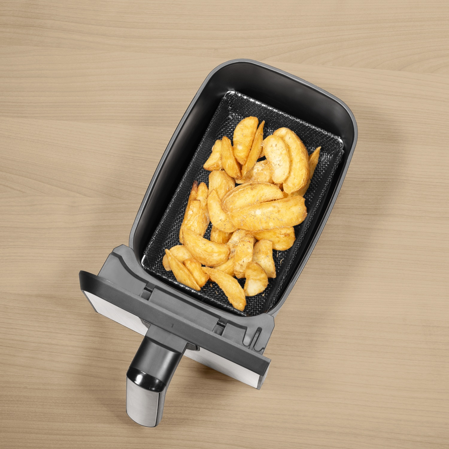An overhead shot of an air fryer with golden potato wedges inside. It's on a light wooden surface.