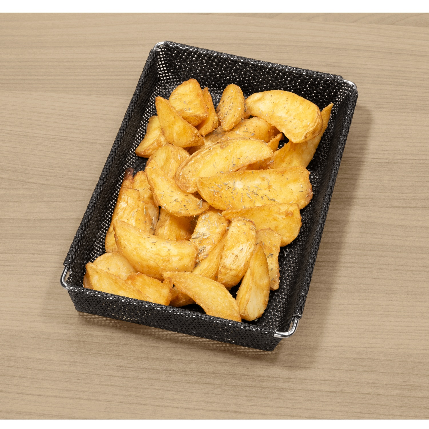 Golden potato wedges in a black basket on a wooden table. Seasoning visible. Background is light brown wood grain.