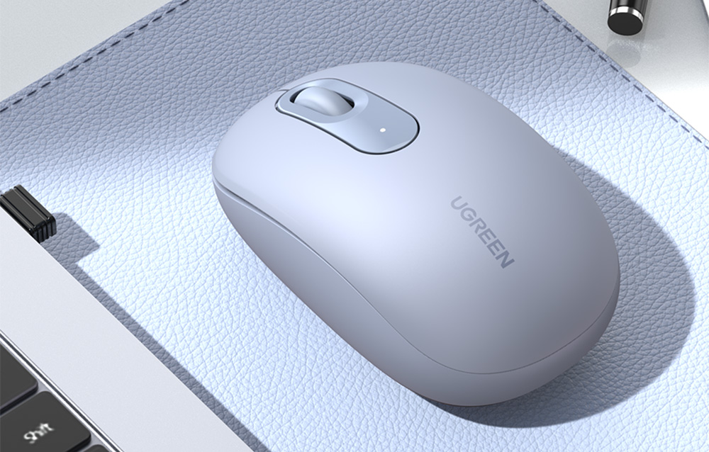 Gri bir UGREEN kablosuz fare, bir dizüstü bilgisayarın ve bir kalemin yanında, açık mavi deri bir mousepad üzerinde duruyor.