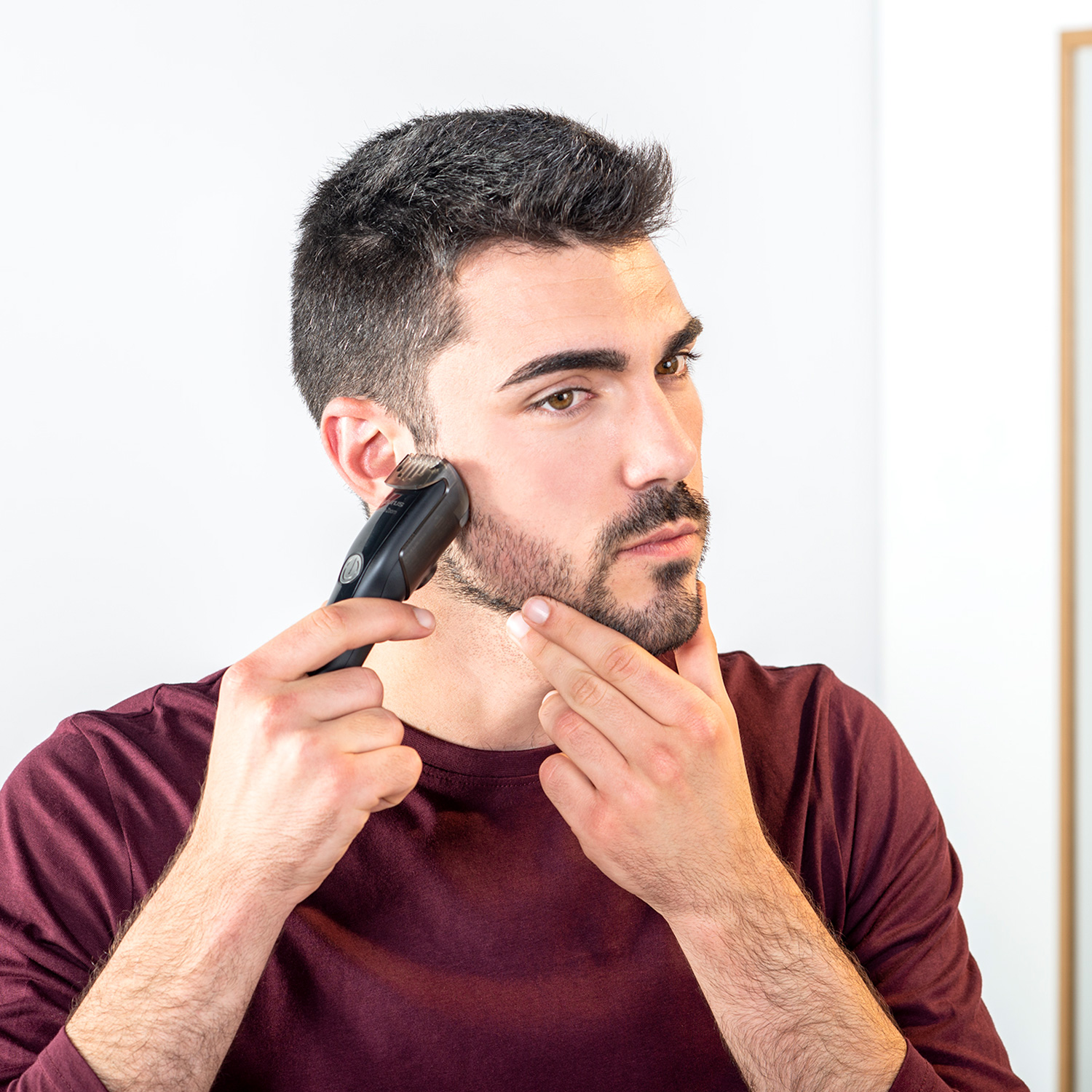 Un hombre se afeita la barba con una recortadora eléctrica. Sostiene la recortadora en su mejilla, mirándose en un espejo. Lleva una camisa granate.
