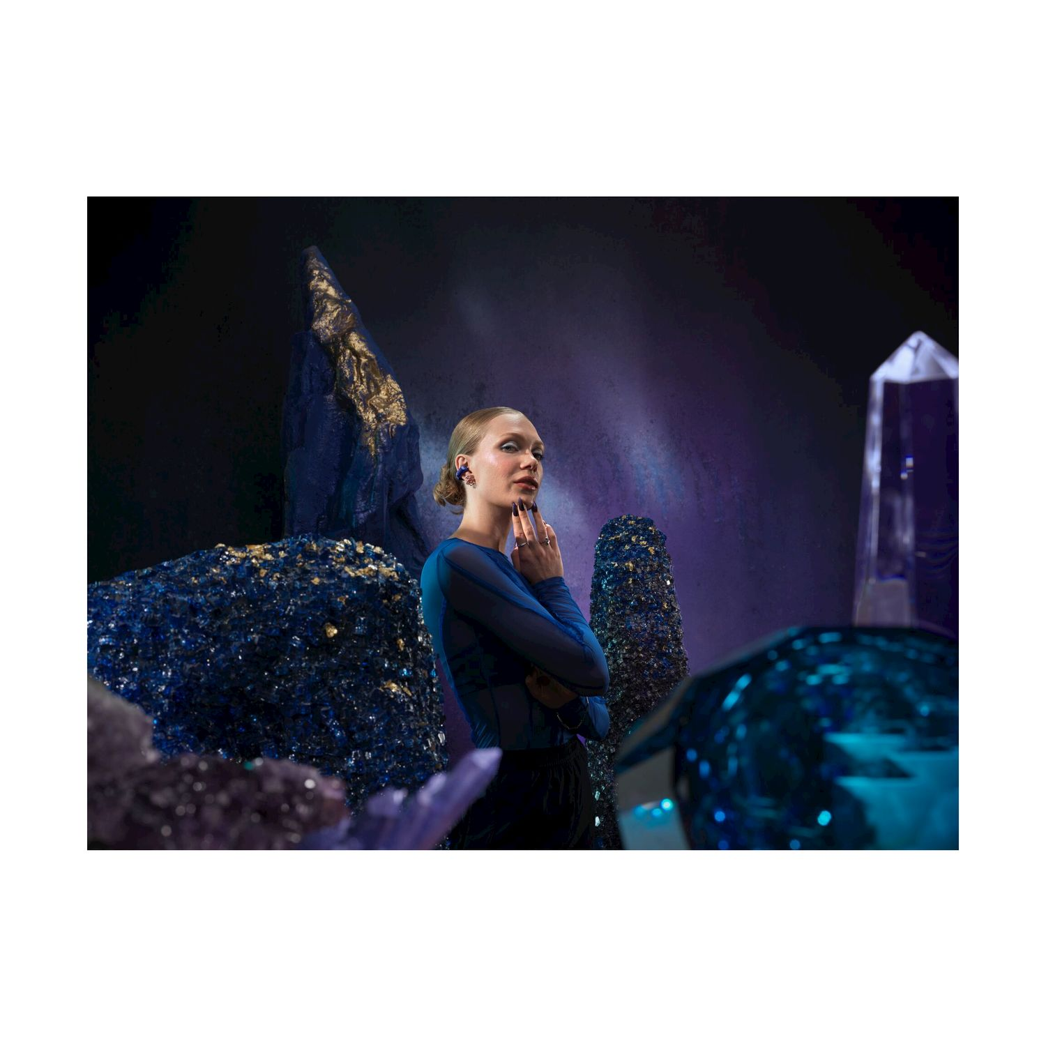 Donna con top blu, circondata da cristalli. Indossa orecchini. Sfondo scuro con elementi blu e oro.