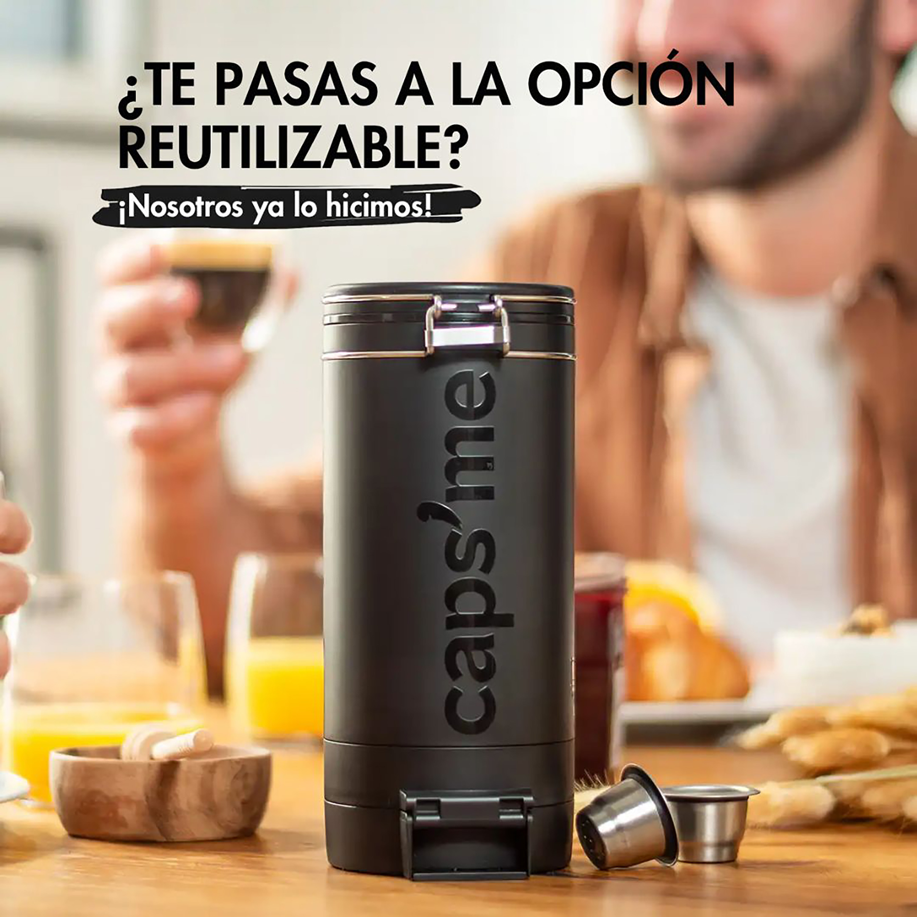 Una cápsula de café reutilizable negra con un cierre plateado está sobre una mesa. Una persona sostiene una taza de café y hay desayuno.