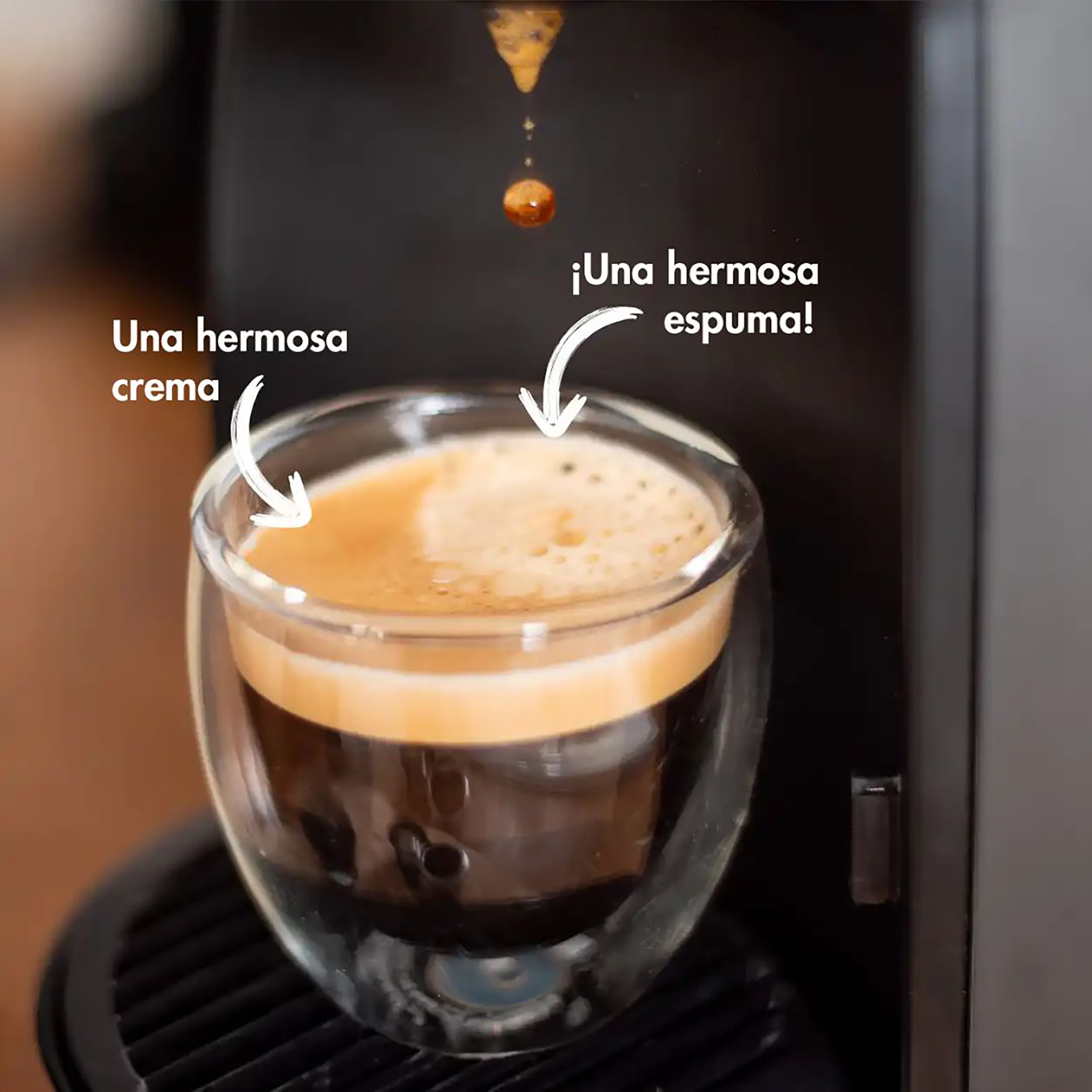 Un vaso de espresso con crema y espuma, una gota de café y texto en español. Cafetera al fondo.