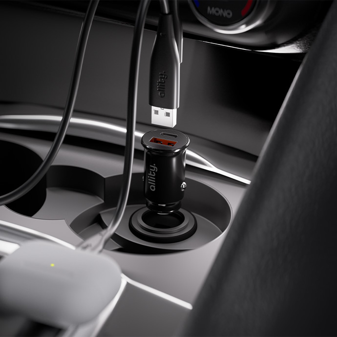 Egy fekete autós töltő, USB kábellel. Egy fehér töltőtok van előtérben. A töltő egy autós pohártartóban van.