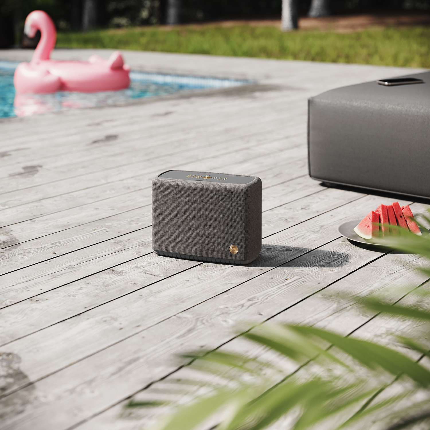 A speaker on a wooden deck next to a pool. A pink flamingo float, a gray ottoman, and watermelon slices are visible.