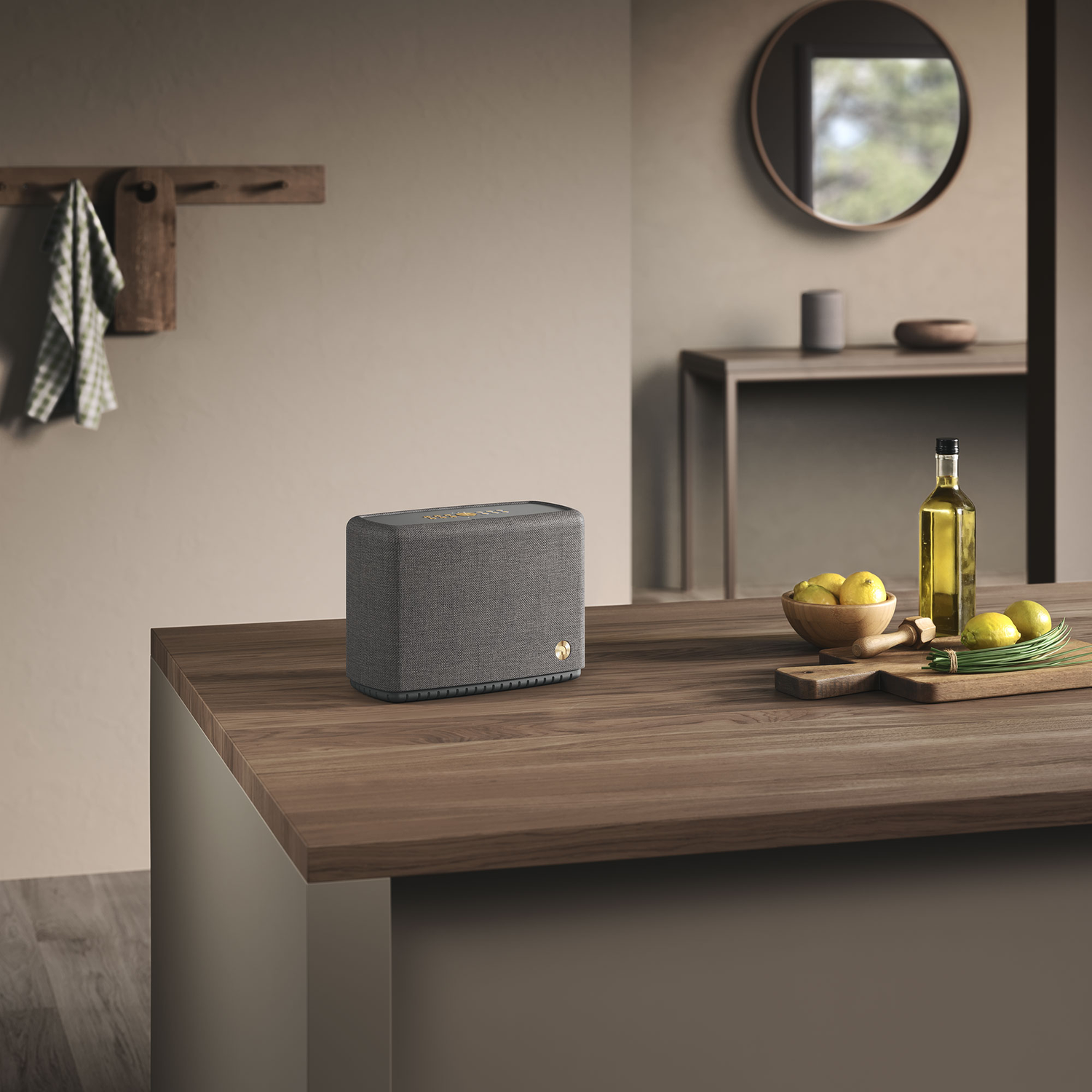 A gray speaker on a wooden counter with lemons, oil bottle, and a mirror in the background. A towel hangs nearby.