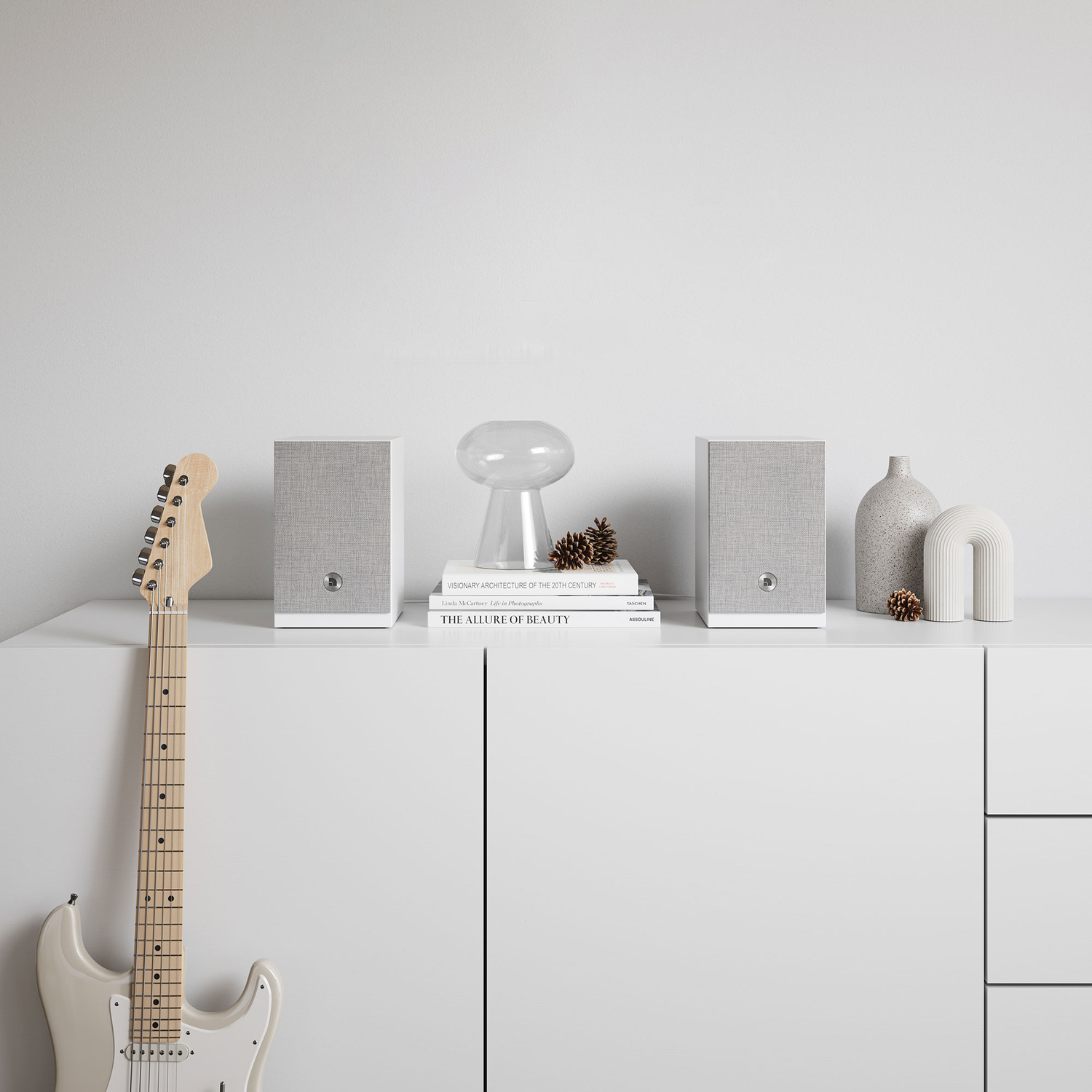 Een witte kast met luidsprekers, boeken en decoratieve items. Een gitaar leunt tegen de kast. De achtergrond is een witte muur.