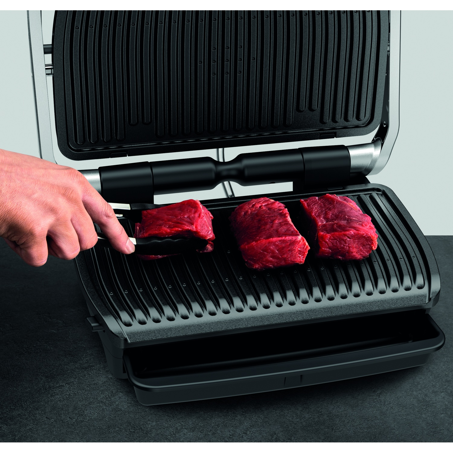 Un grill avec trois steaks crus. Une main utilise des pinces pour en manipuler un. Le grill est noir, le fond est gris clair.