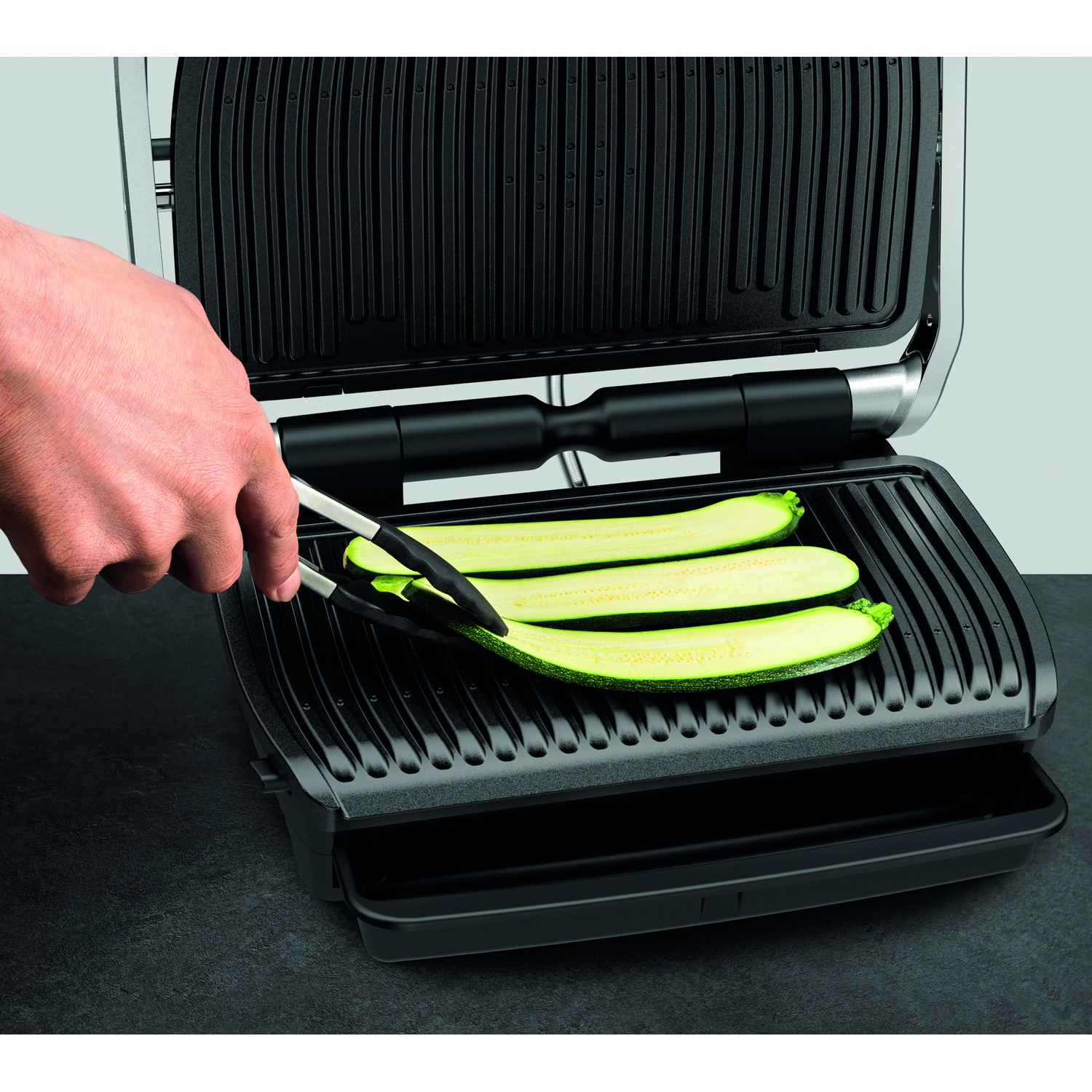 Une main utilise des pinces pour placer des tranches de courgettes sur un grill noir. Le grill est ouvert, et le fond est un mur uni.