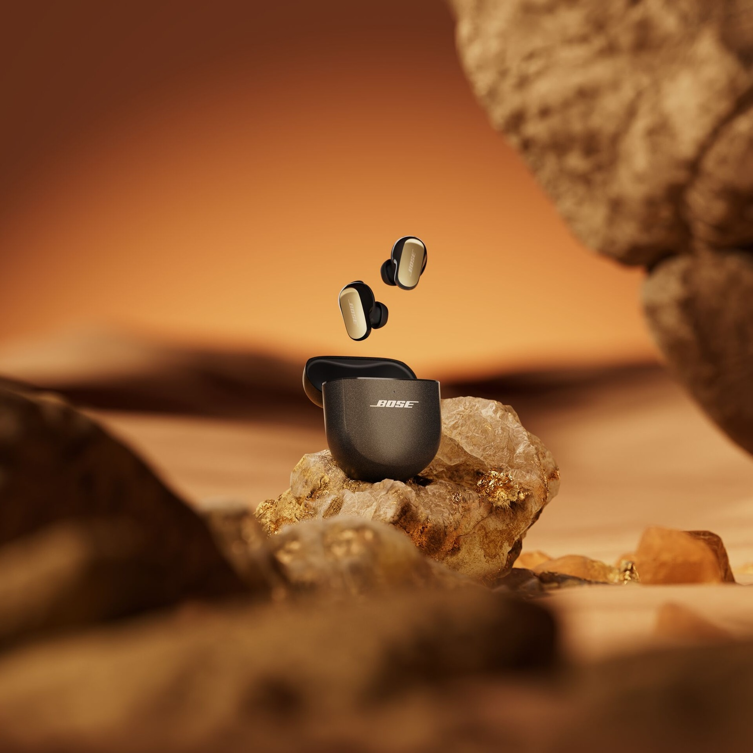 Auricolari Bose che fluttuano sopra una custodia di ricarica su una roccia. Gli auricolari sono neri e oro, la custodia è nera. Lo sfondo è arancione.