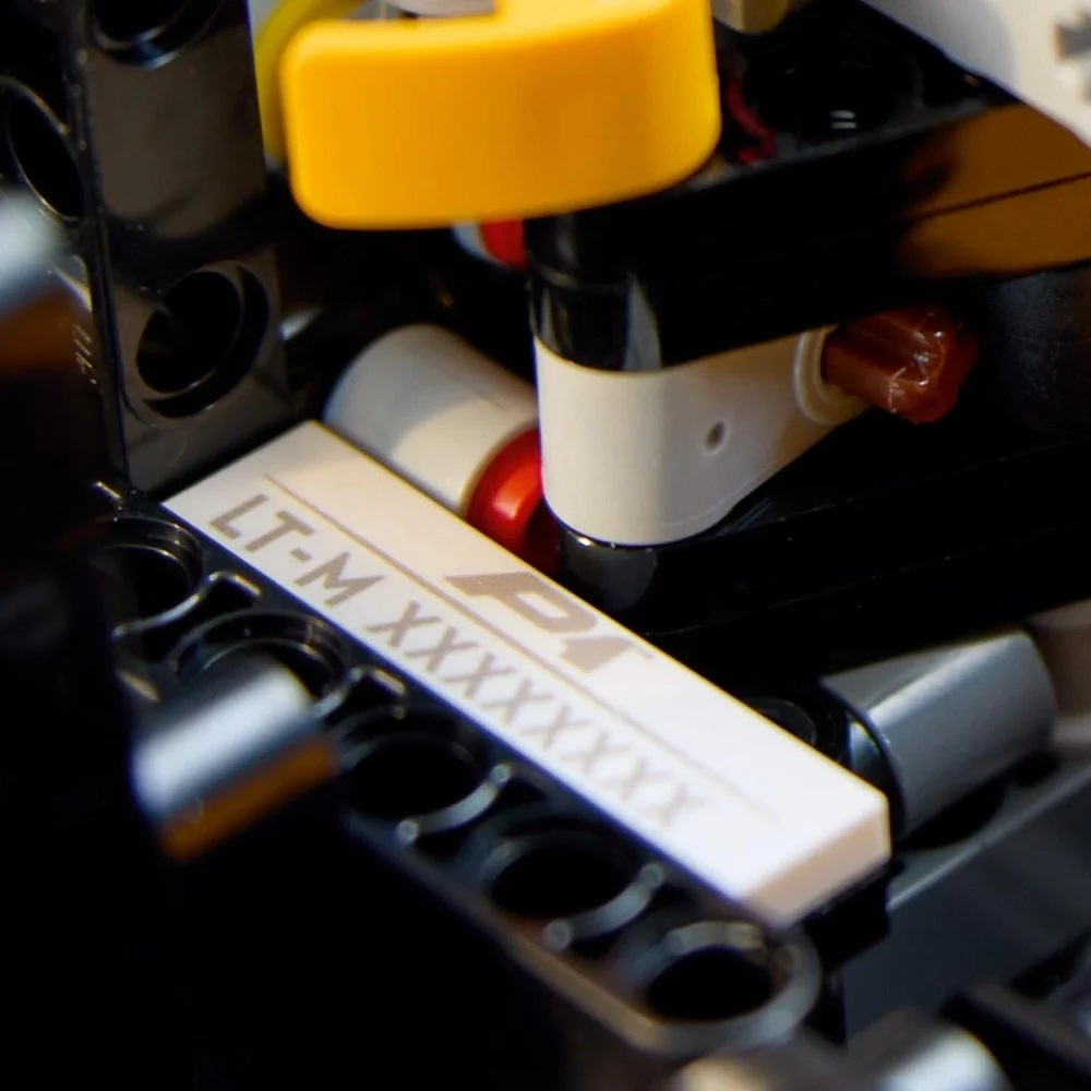 Zbliżenie białej klocki Lego z tekstem i logo. Inne części Lego są czarne, białe, czerwone i żółte, zmontowane w maszynie.