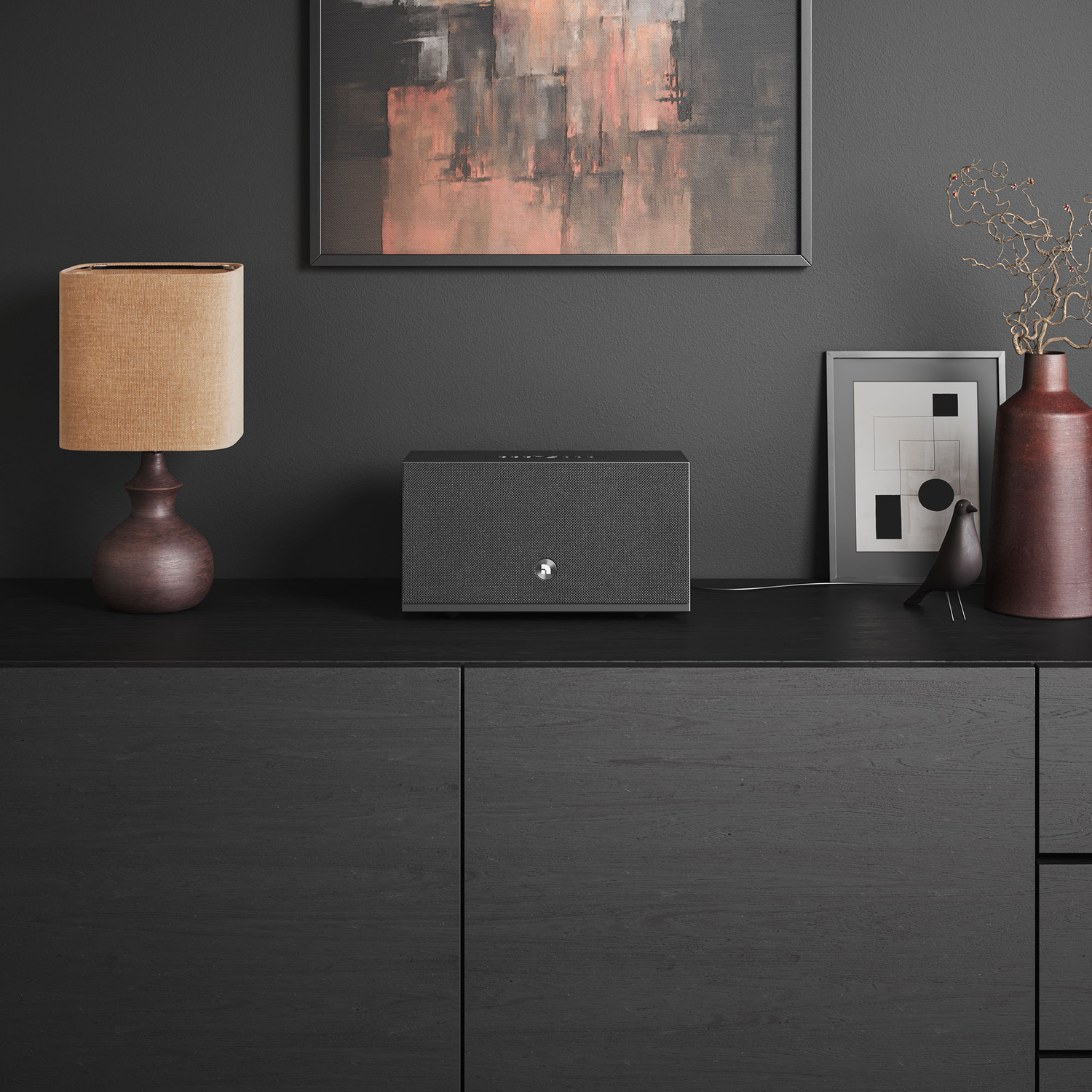 Een zwarte kast met een luidspreker, lamp, kunstwerk en decor. De achtergrond is een donkergrijze muur.