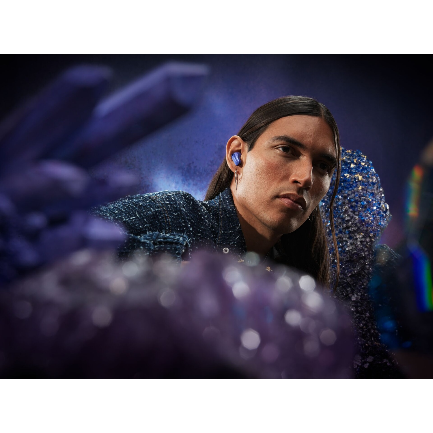 Un uomo con i capelli lunghi indossa una giacca blu e un auricolare blu. Cristalli viola in primo piano e sullo sfondo.