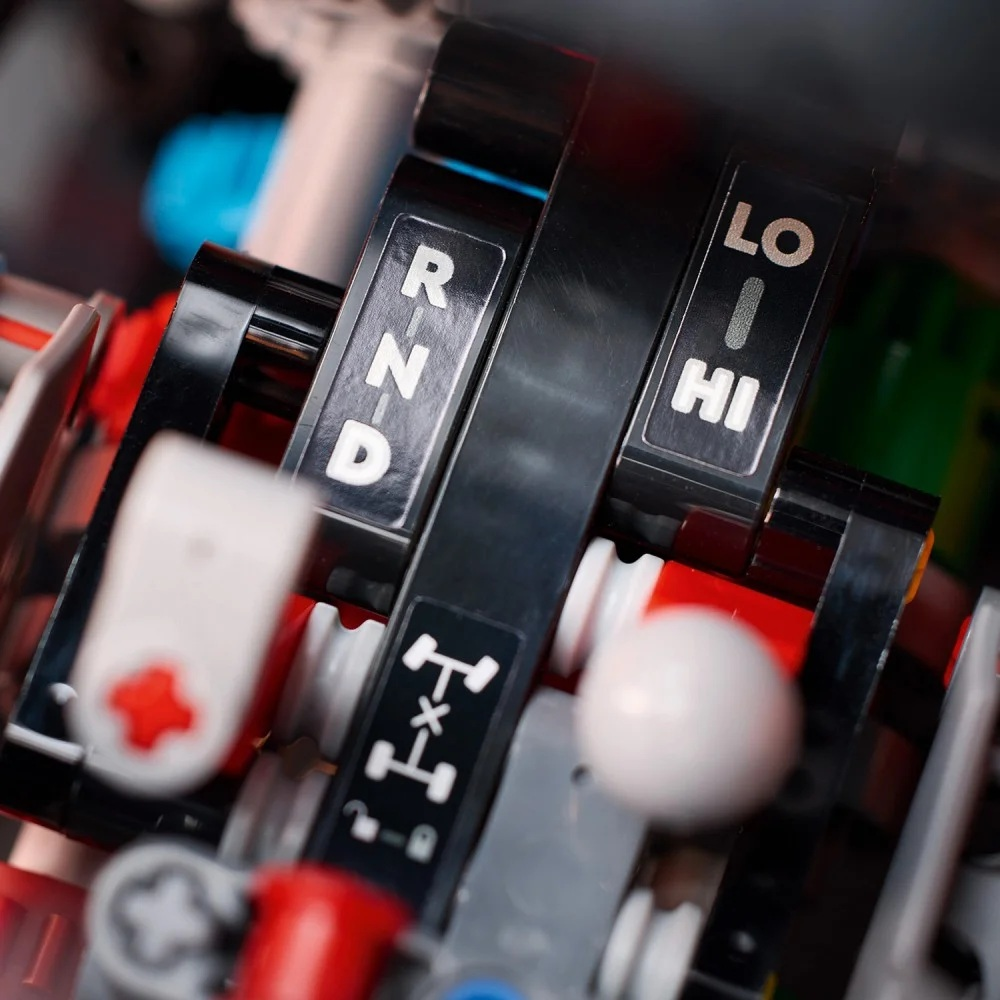 Zbliżenie czarnej dźwigni zmiany biegów z białymi literami: R, N, D, LO, HI. Widoczne są czerwone, białe i szare elementy.