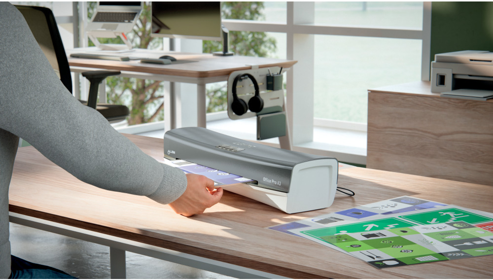 Egy személy egy 'Office Pro A3' feliratú laminálógépet használ egy faasztalon. A háttérben egy íróasztal és egy ablak látható.