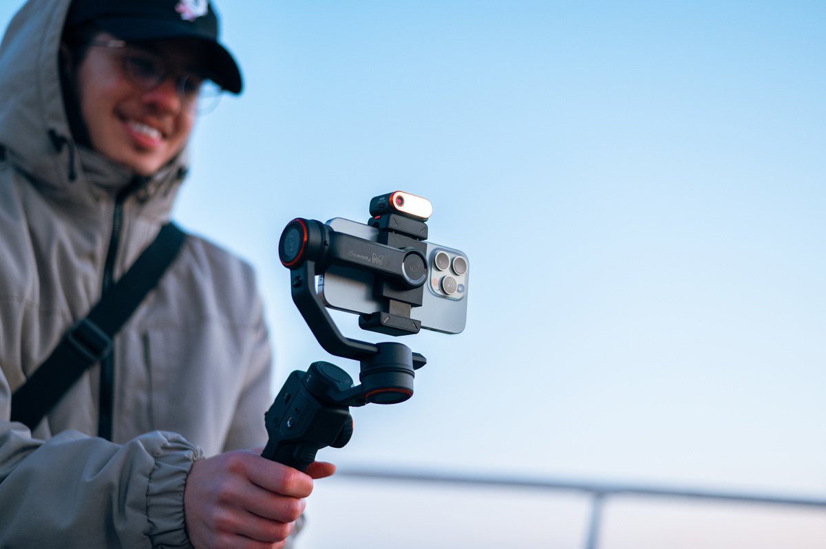 Eine Person hält ein Telefon auf einem Gimbal und lächelt. Der Hintergrund ist ein klarer blauer Himmel.