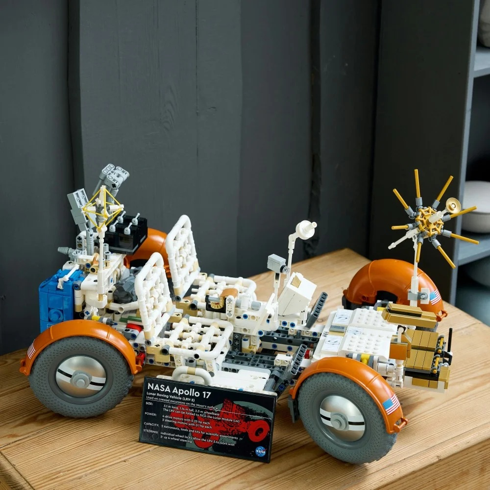 Model Lego NASA Apollo 17 Lunar Roving Vehicle stoi na drewnianej powierzchni. Jest biały, pomarańczowy i szary, z czarną kartą informacyjną.