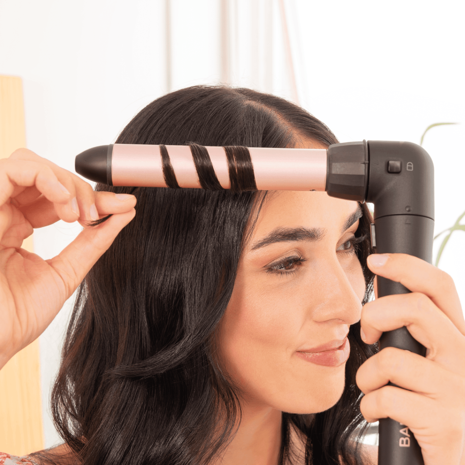 Una mujer se riza el pelo oscuro con una plancha. La plancha es oro rosa y negra. Ella está mirando su cabello.