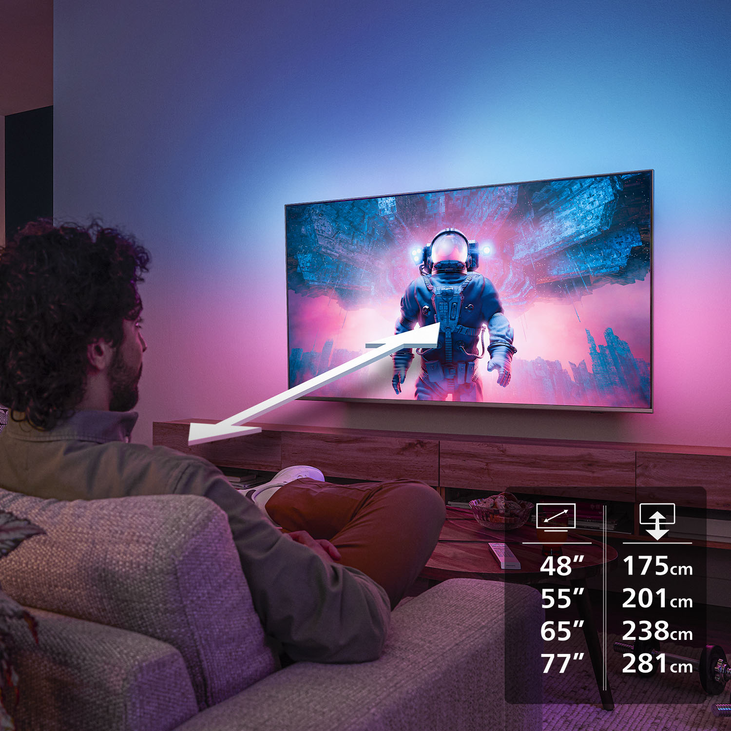 Un hombre mira un televisor con un astronauta. Se muestran los tamaños del televisor. La habitación tiene iluminación ambiental.