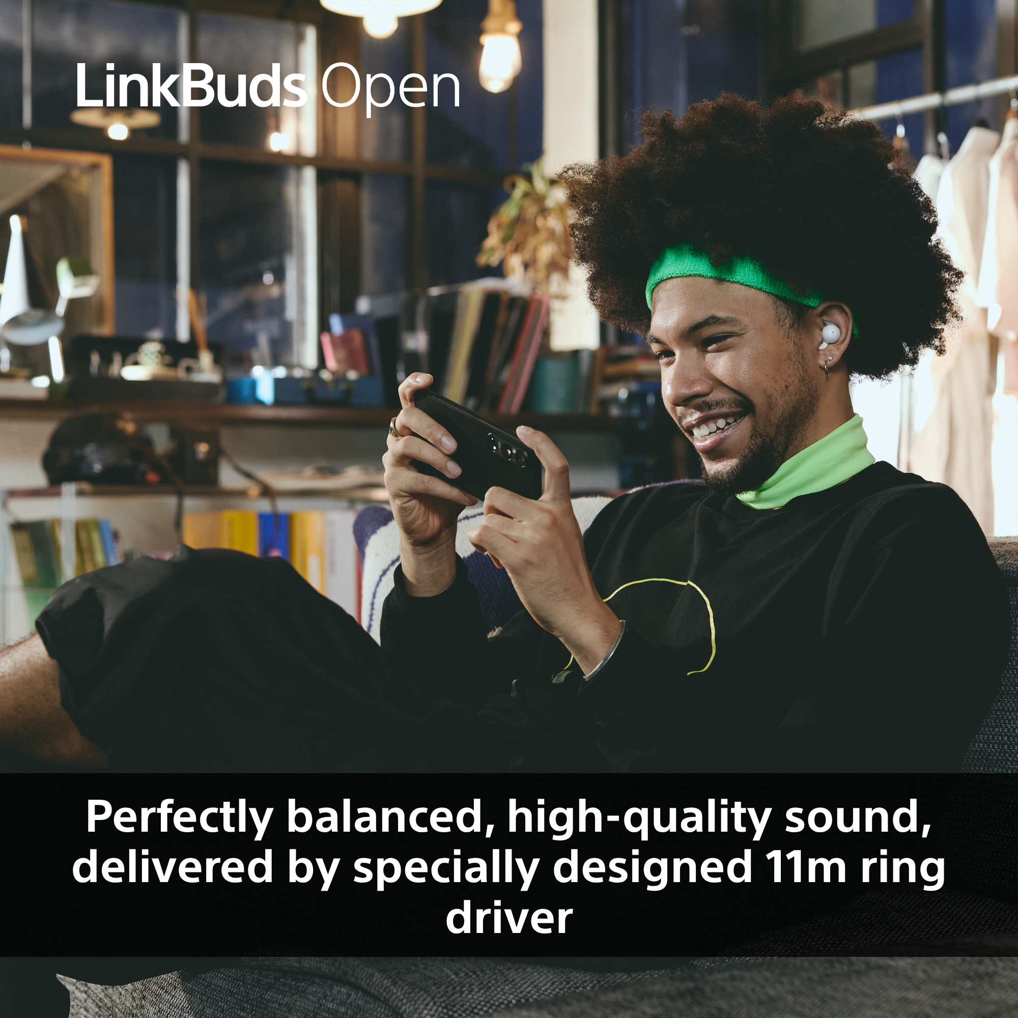 Un homme avec des écouteurs tient un téléphone, souriant. Le fond a une fenêtre, un portant et un texte qui dit 'LinkBuds Open'.