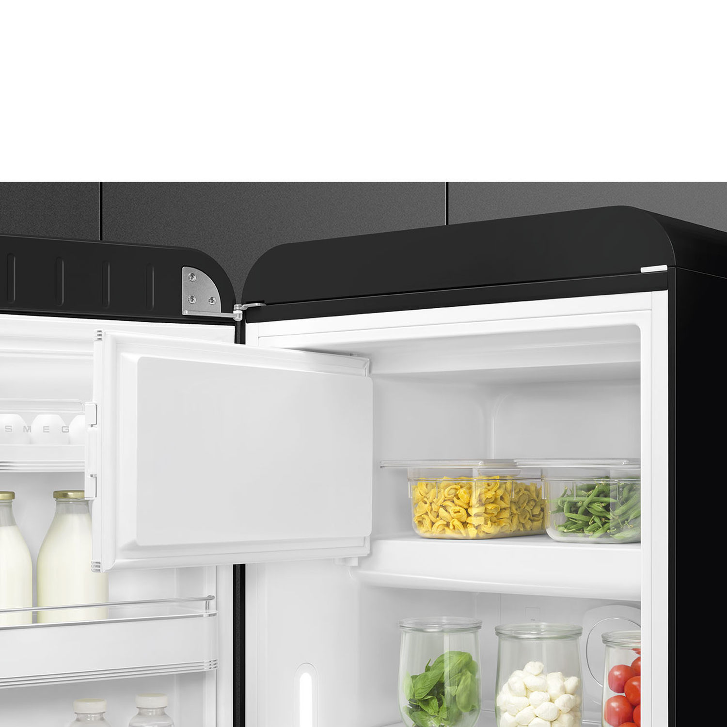Open black refrigerator. Inside, containers with pasta, green beans, basil, mozzarella, and tomatoes are visible.